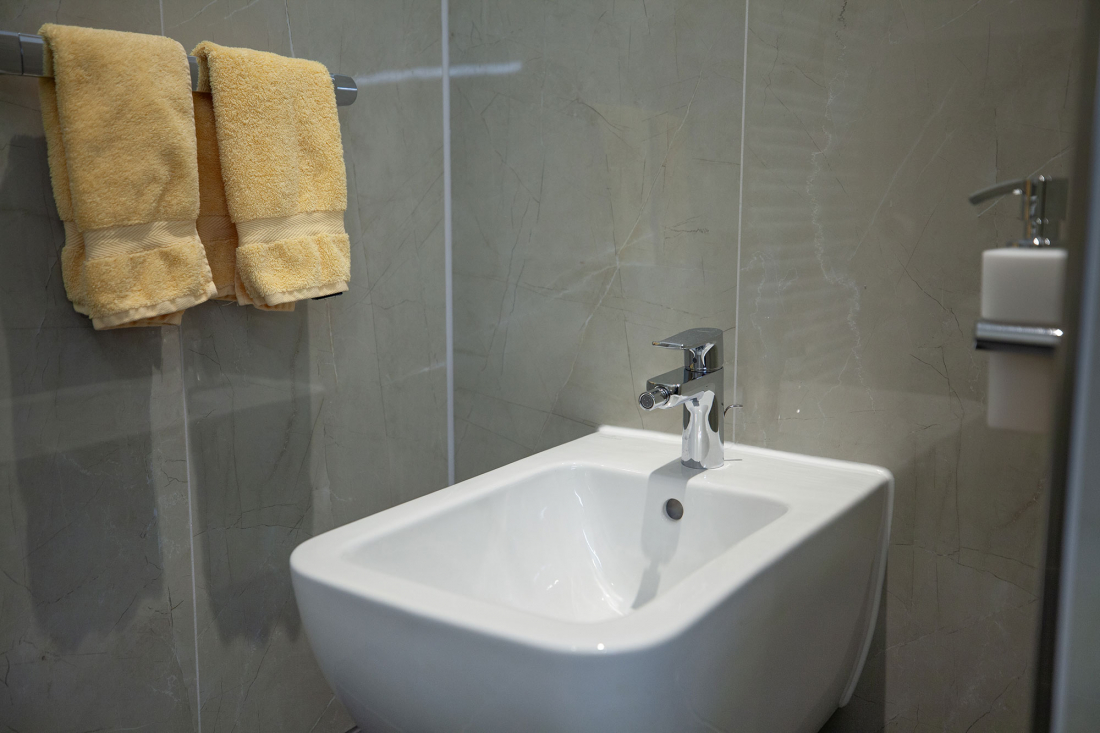 Ein modernes weißes Bidet mit einem verchromten Wasserhahn ist vor einer grau gefliesten Wand installiert. Zwei gelbe Handtücher hängen an einem Handtuchhalter in der Nähe. Rechts an der Wand ist ein Seifenspender angebracht.